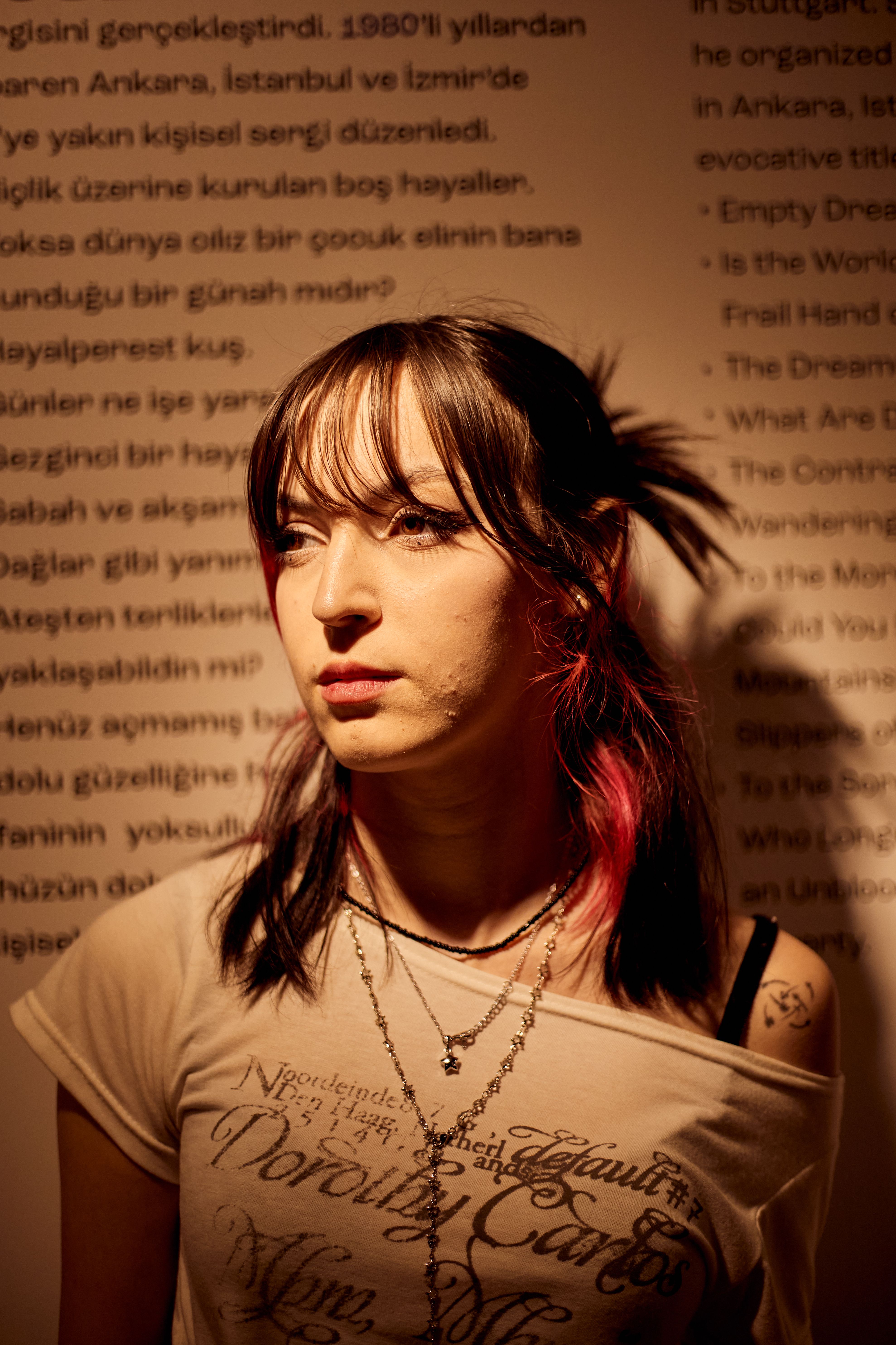 Close-up portrait of a woman with pink-tinted hair standing against text-covered wall in dim lighting.