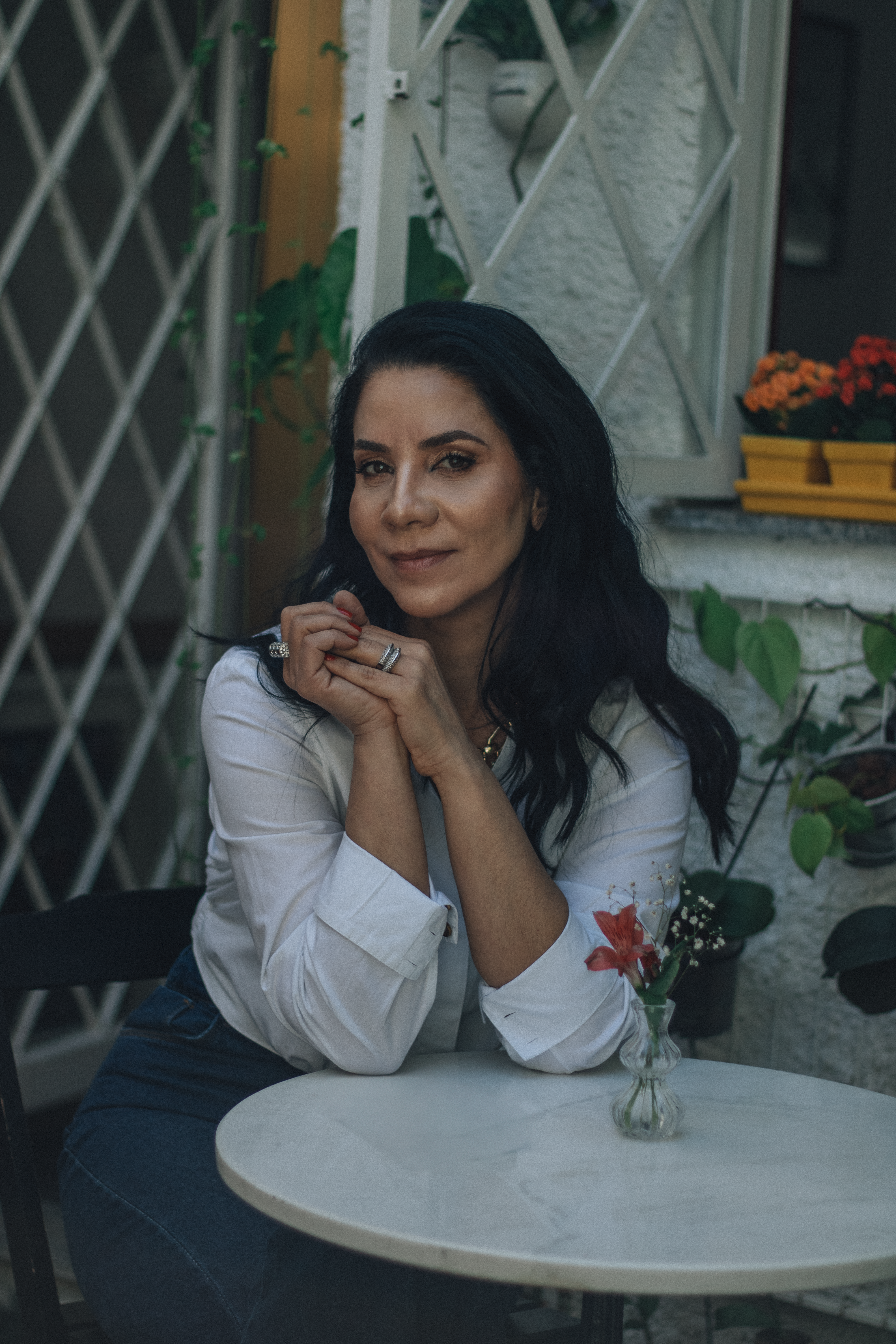 A poised woman seated at a café table outdoors, surrounded by plants.
