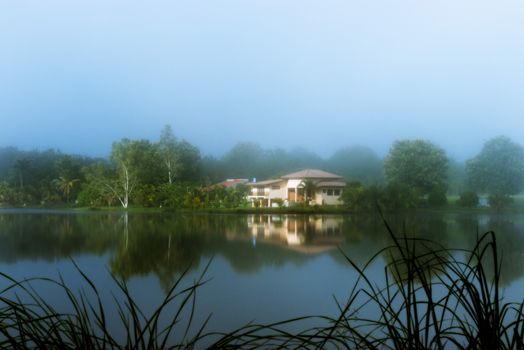 A tranquil view of a house reflected in a misty lake surrounded by lush greenery.