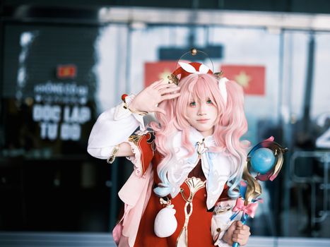 A young adult in elaborate pink cosplay costume poses with a friendly wave.