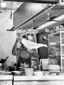 Monochrome image of a chef stretching dough in an Asian street food stall with various kitchen items.