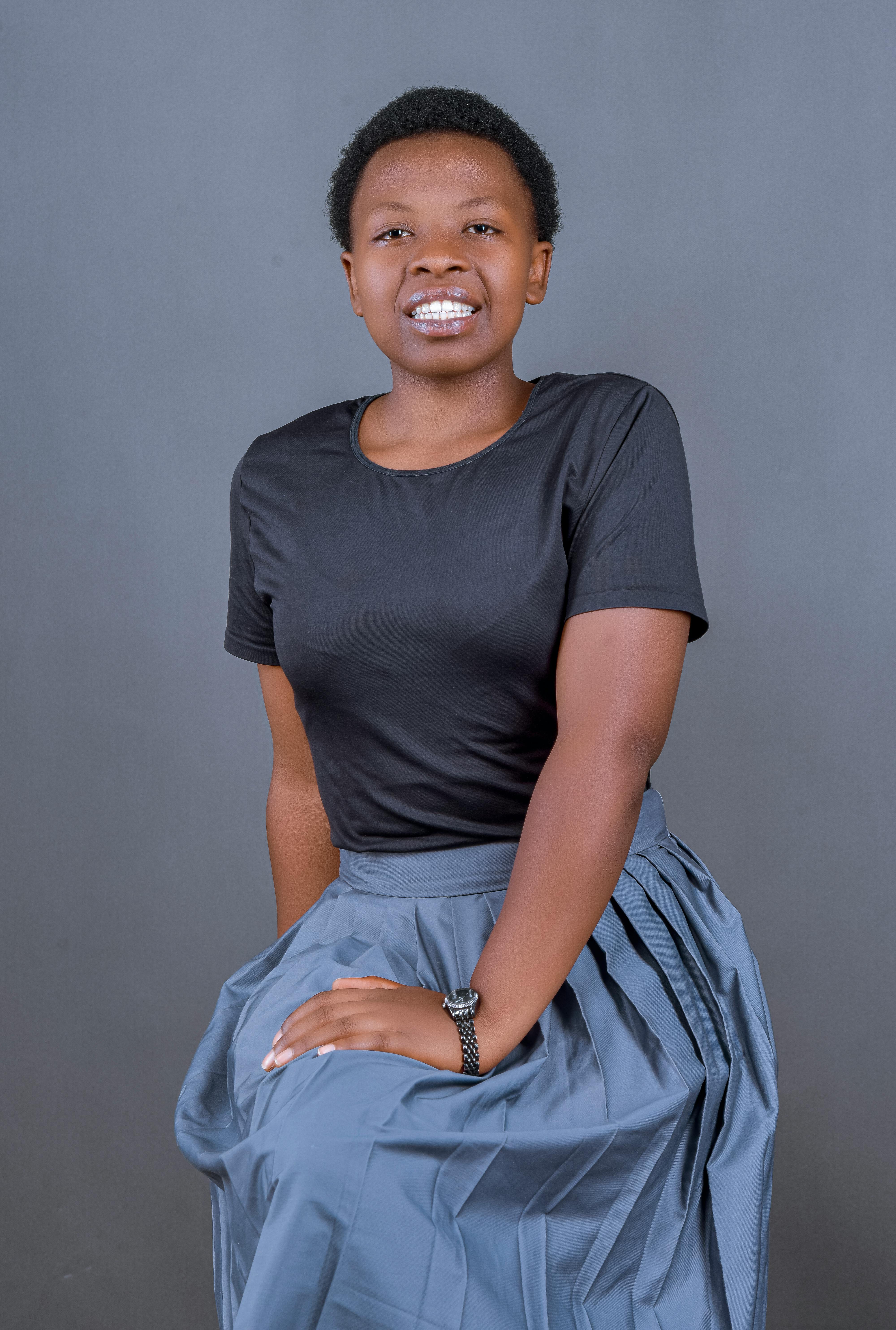 A cheerful woman sitting in a studio wearing a black top and gray skirt, smiling brightly.