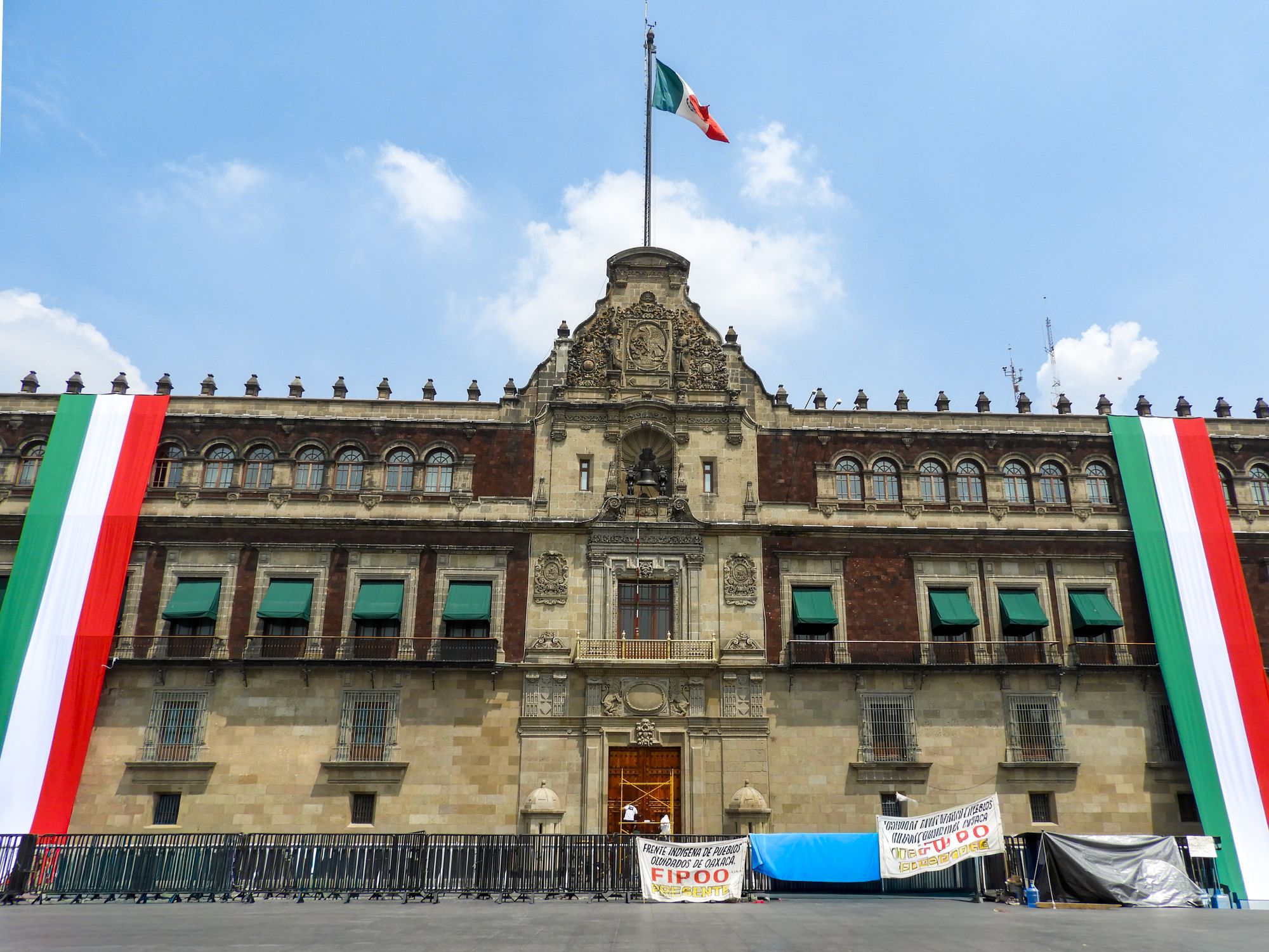 Front view of the Mexican National Palace in Mexico City with large Mexican flags displayed, a bright sky overhead.