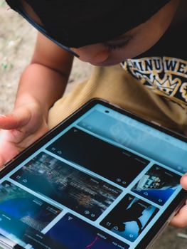 A young child intensely focused on a tablet screen while sitting outdoors, wearing casual attire.