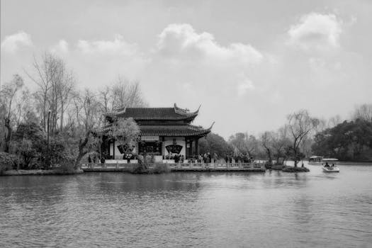 Black and white image of a traditional Chinese pavilion by a serene lake, with trees and boats.