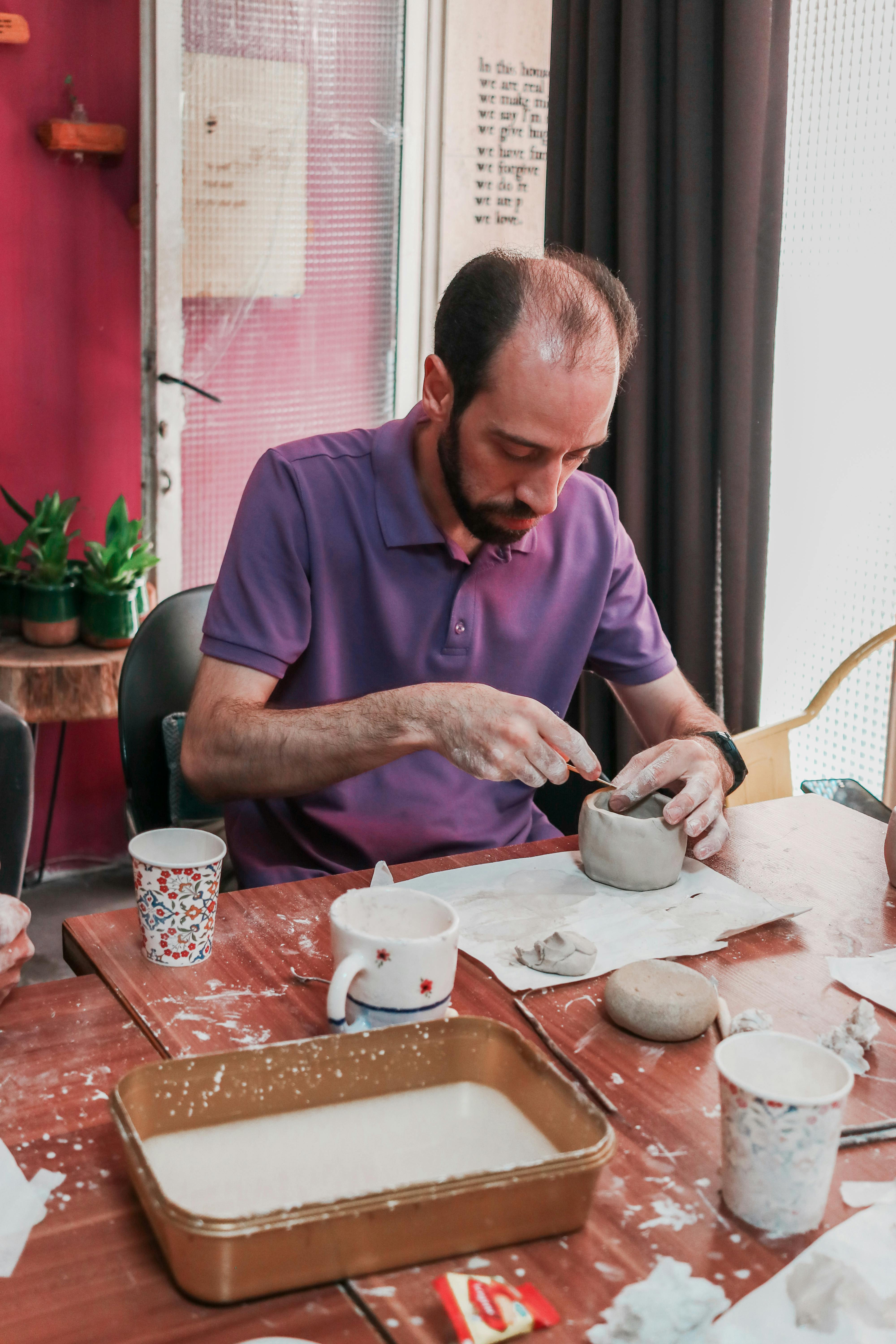 Adult man sculpting clay in a pottery workshop with tools on table.