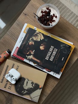 Flat lay of books, yogurt with raisins, earbuds, and a beverage indoors on a wooden table.