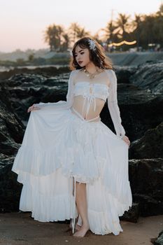 A woman in a flowing white dress poses on a rocky beach at sunset.