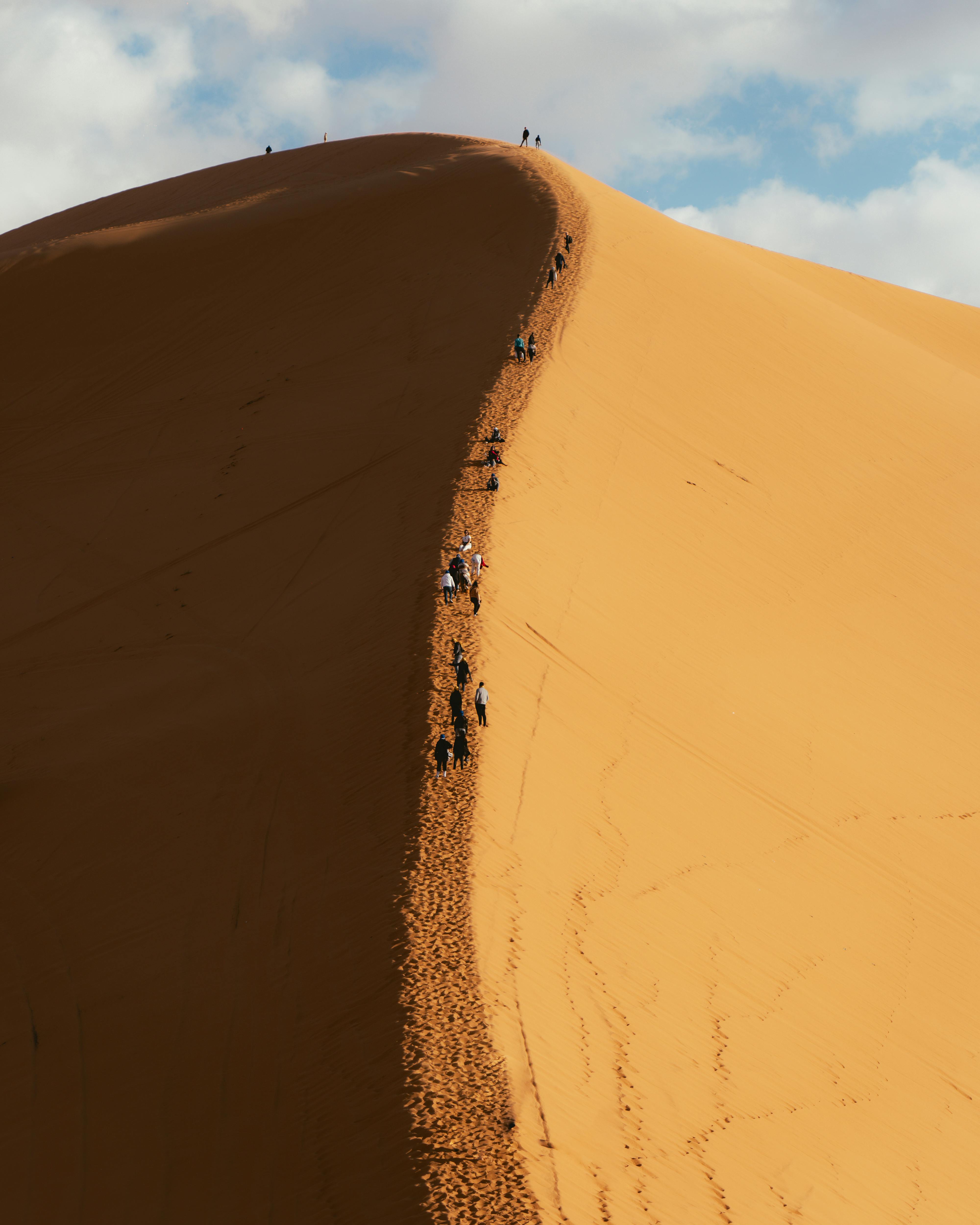 A group of people climbing a large sand dune under a bright sky, showcasing adventure and nature's beauty.
