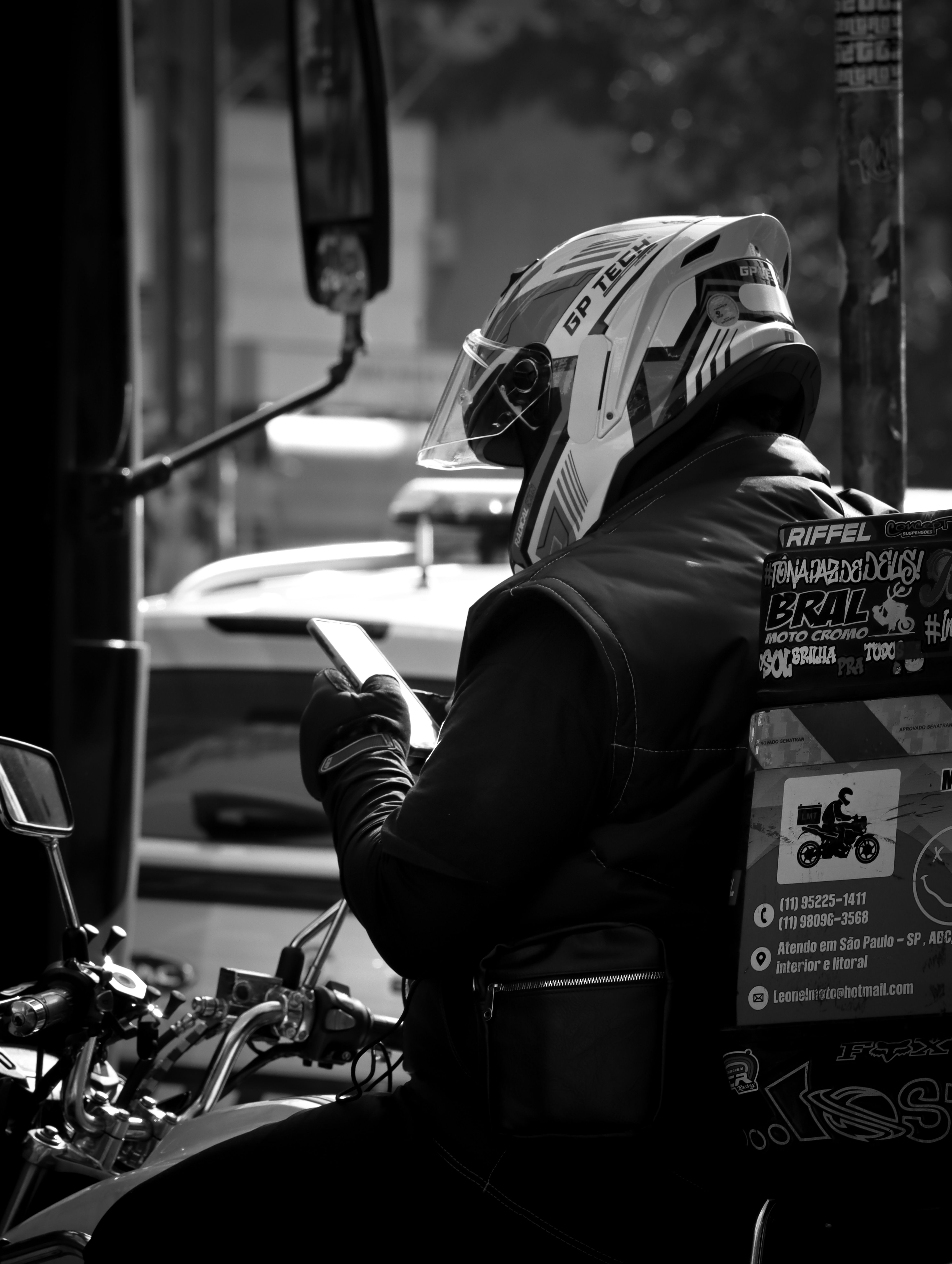 Black and white photo of a delivery rider on a motorcycle using a smartphone in urban setup.