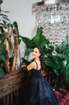 Woman in a sparkling black gown posing elegantly with tropical plants and a chandelier.