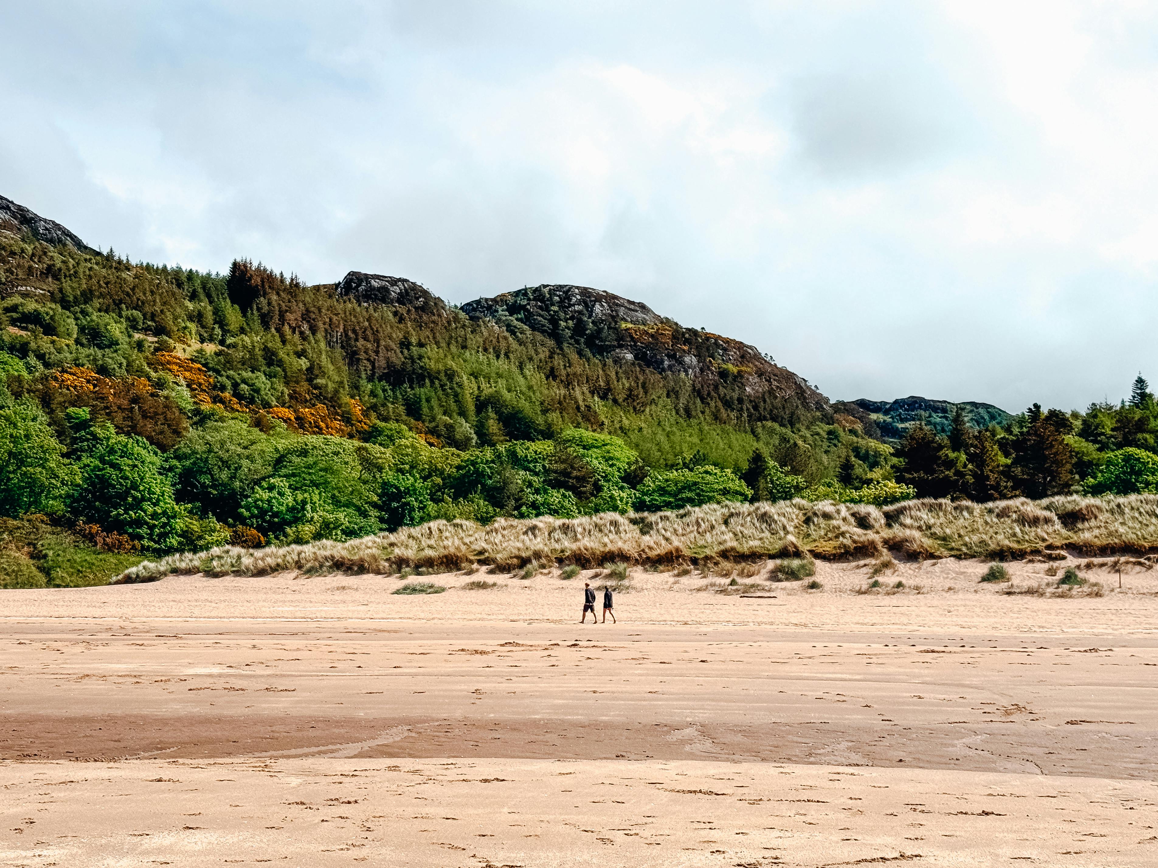 Two people walking on a picturesque beach in Scotland with lush green hills in the background.