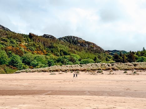 Two people walking on a picturesque beach in Scotland with lush green hills in the background.