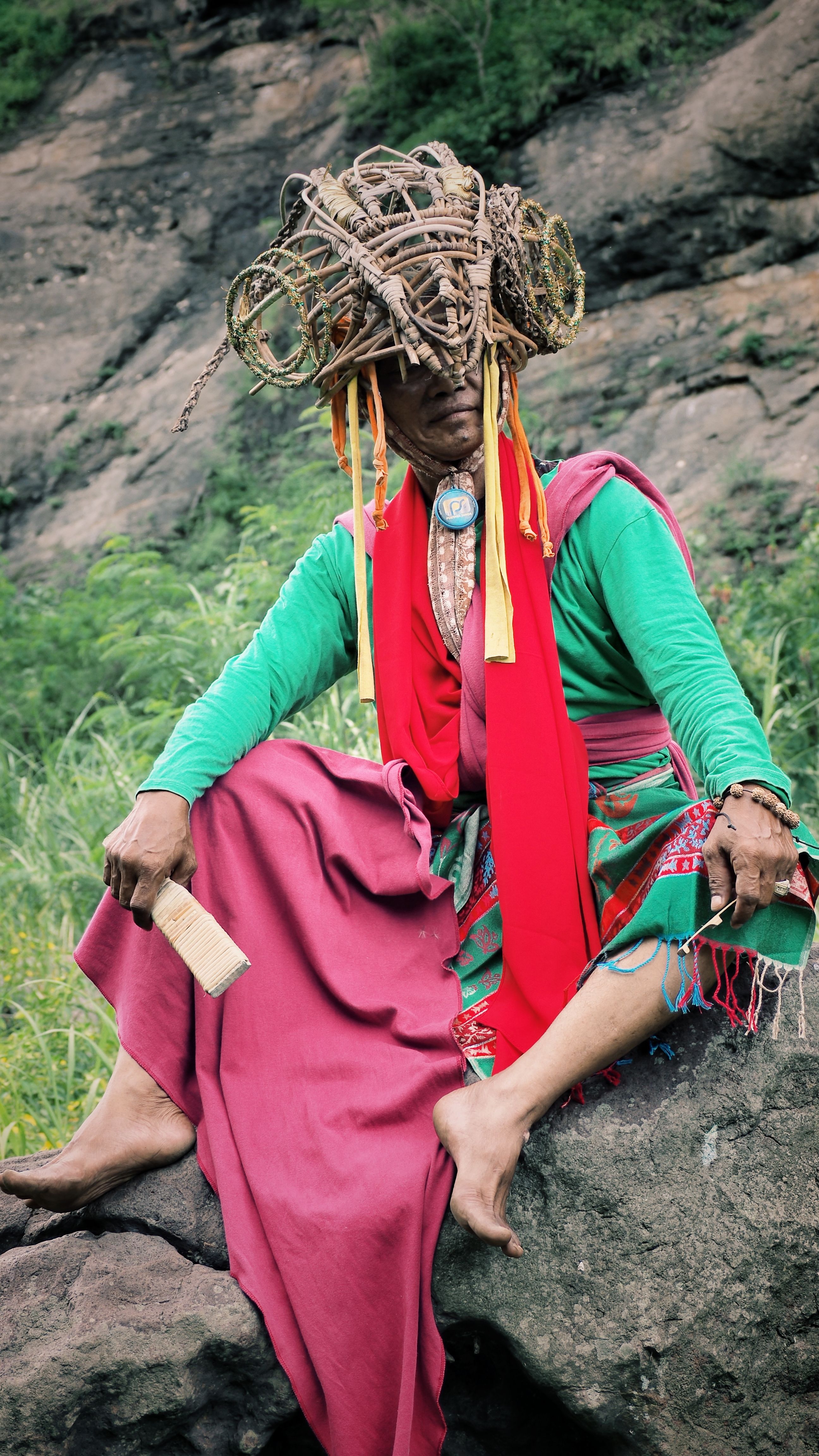Person in vibrant traditional attire with natural headdress sitting outdoors on rocks.
