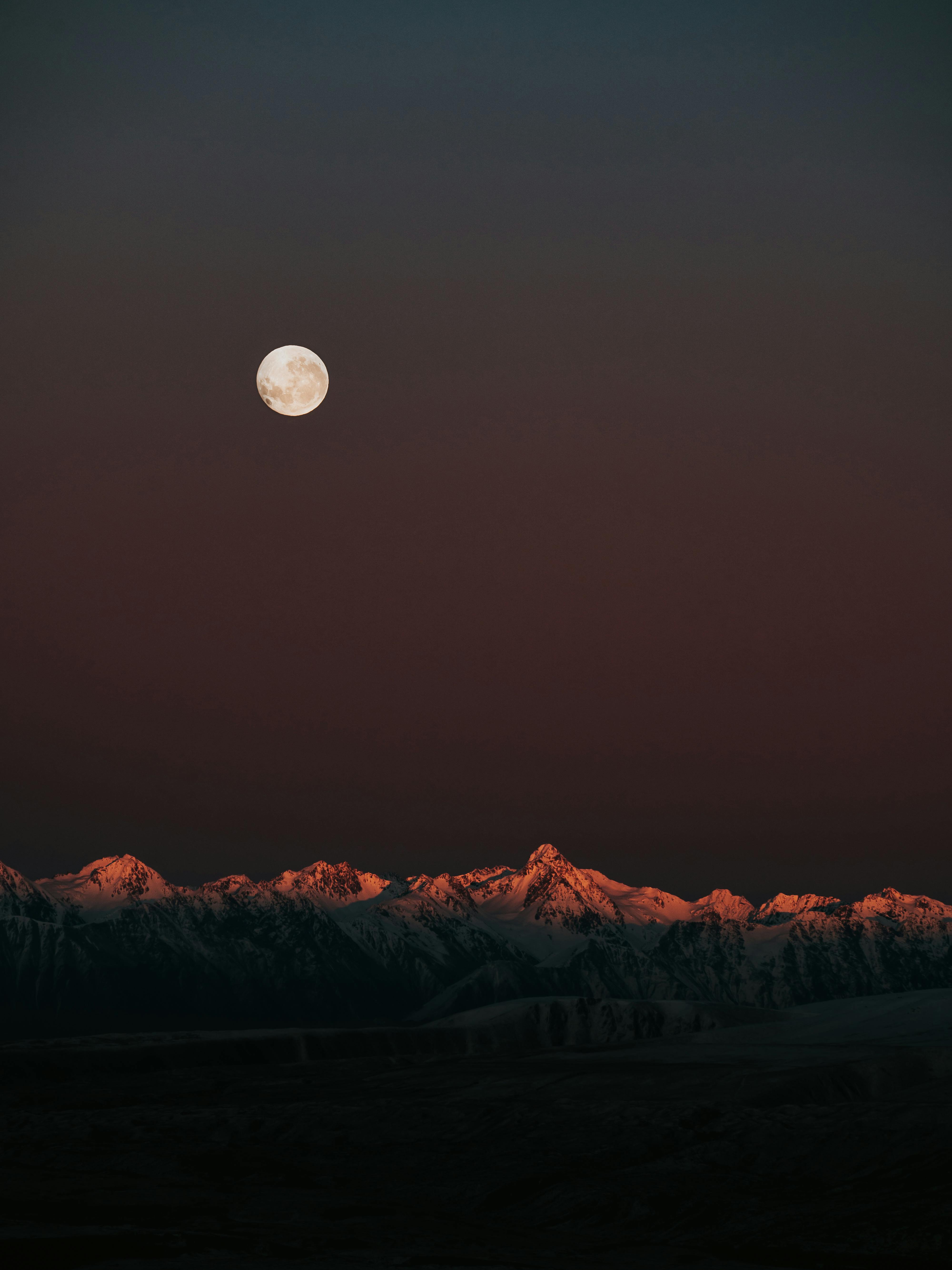 Captivating view of a full moon above snow-capped mountains during twilight.