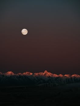 Captivating view of a full moon above snow-capped mountains during twilight.