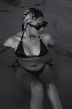 A woman in a bikini and sunglasses relaxes in a pool, captured in monochrome.
