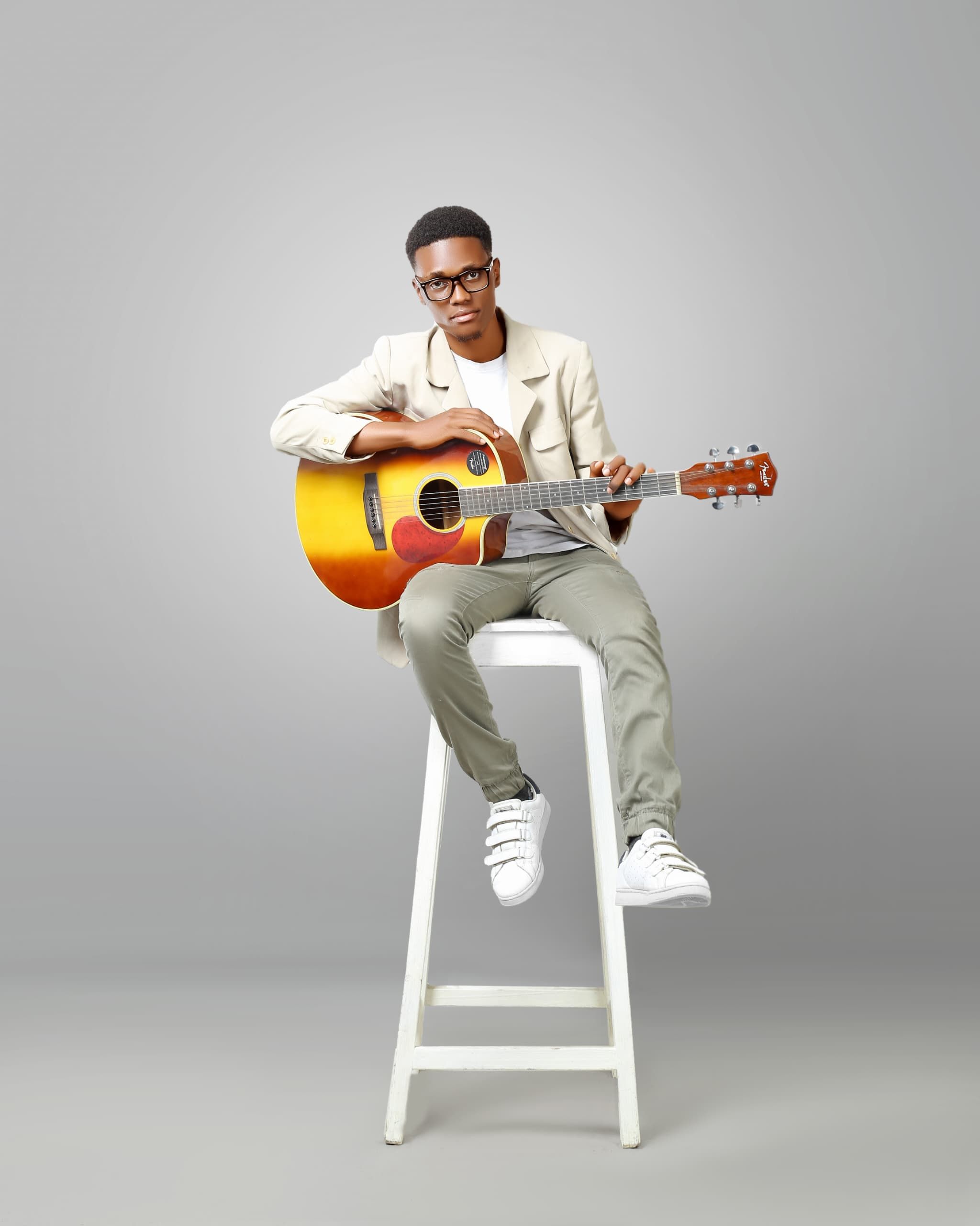 Stylish young adult with an acoustic guitar sitting on a high stool in a minimalist setting.