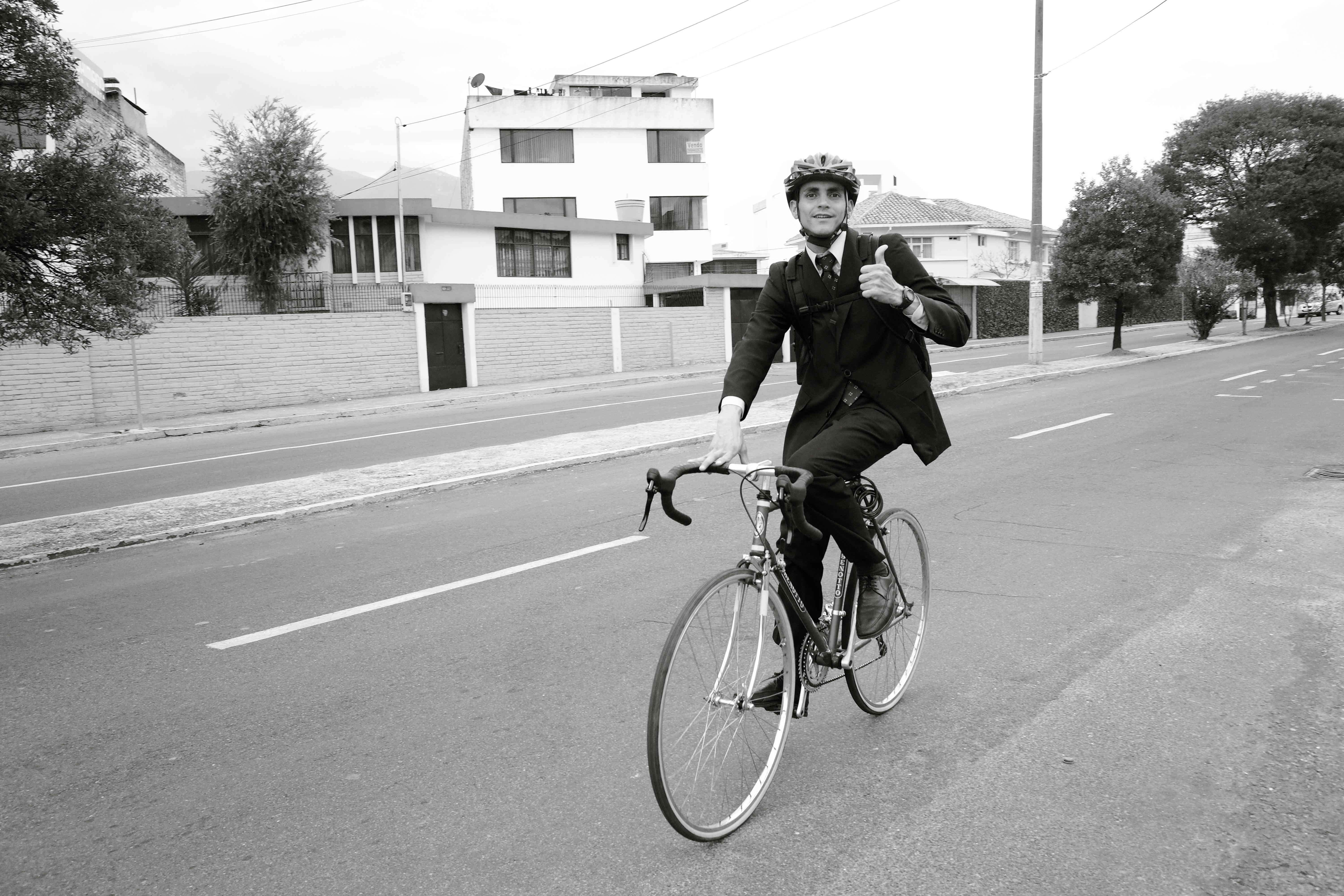Gratis Un uomo d'affari in giacca e cravatta che percorre una strada cittadina in bicicletta, facendo un cenno di saluto con il pollice in su. Foto a disposizione