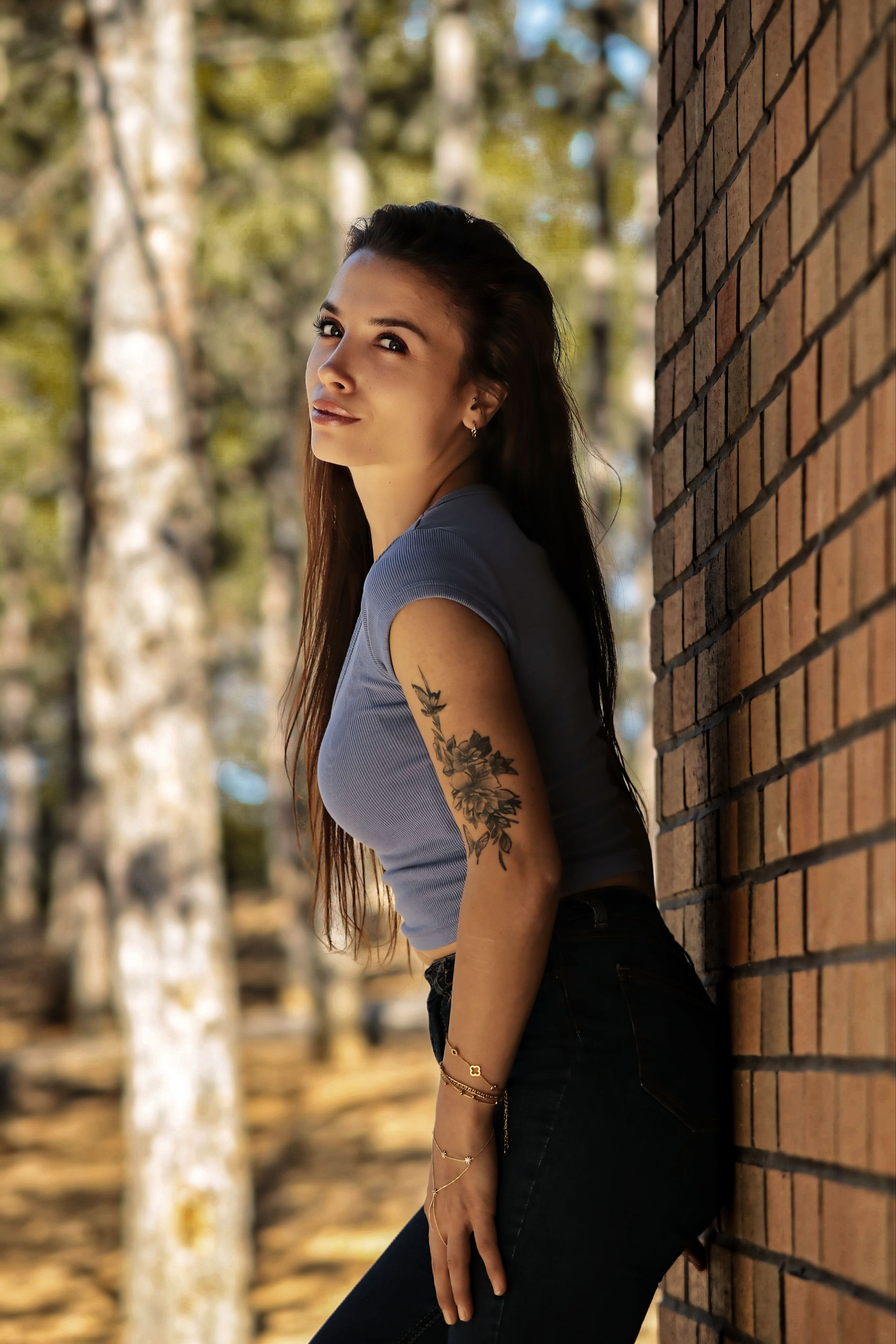 A woman with a tattoo leans against a brick wall in a sunlit forest setting.