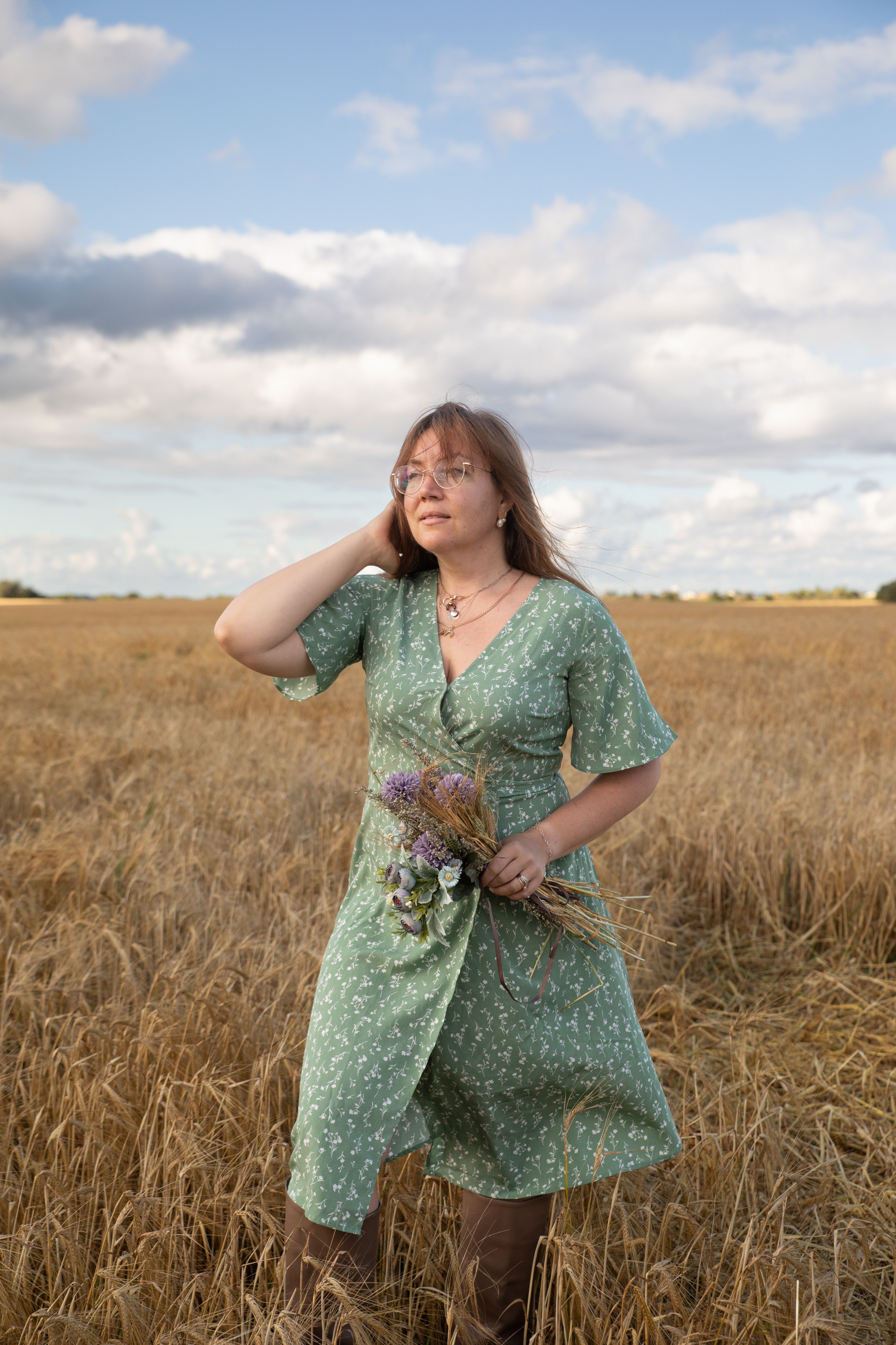 Woman in green dress with flowers standing in golden wheat field under a cloudy sky.