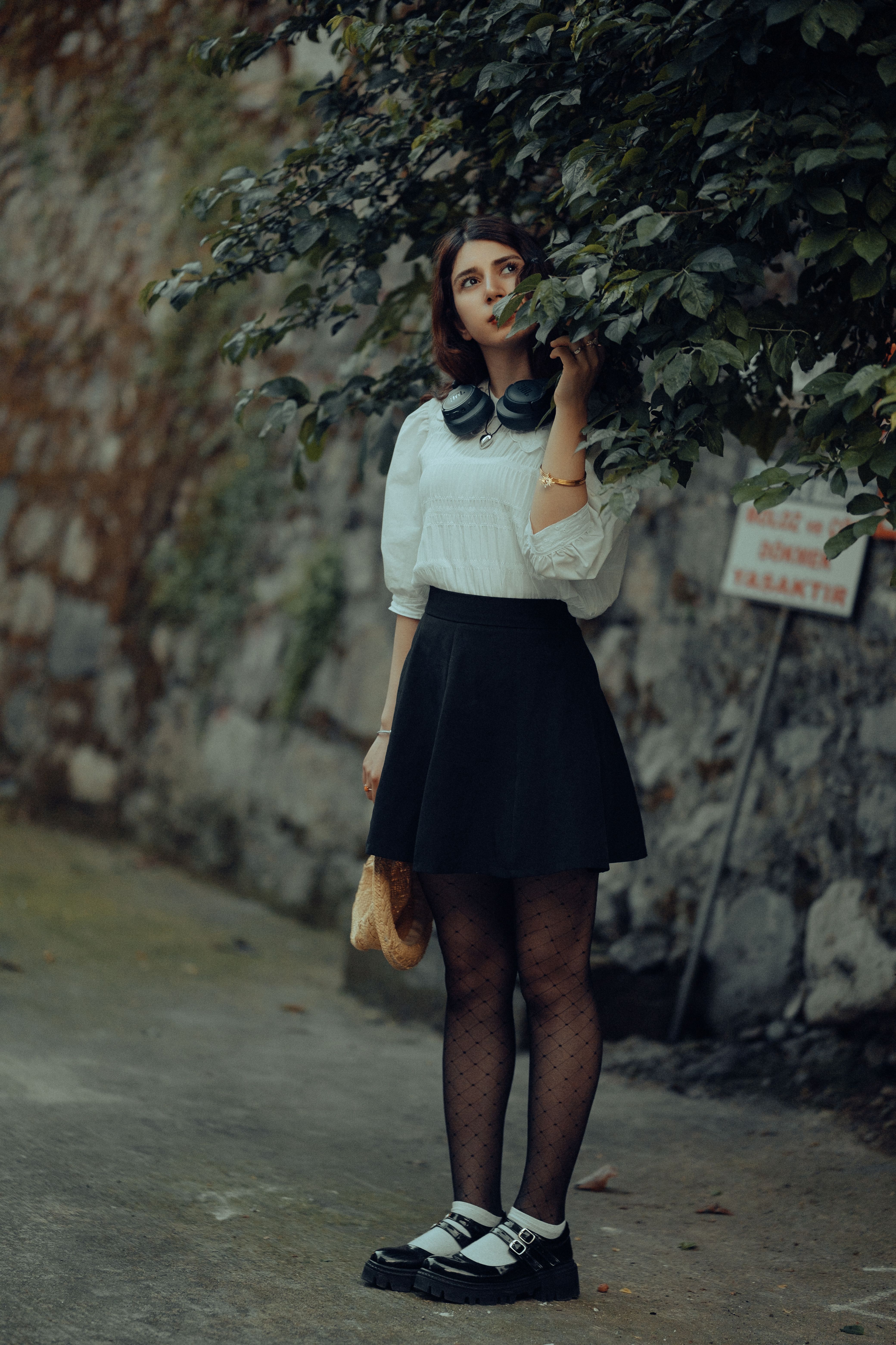 Young woman in chic outfit poses outdoors with a leafy background, exuding modern urban style.