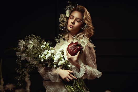 A woman holds a bouquet and a heart in a dark, artistic setting, conveying elegance and emotion.