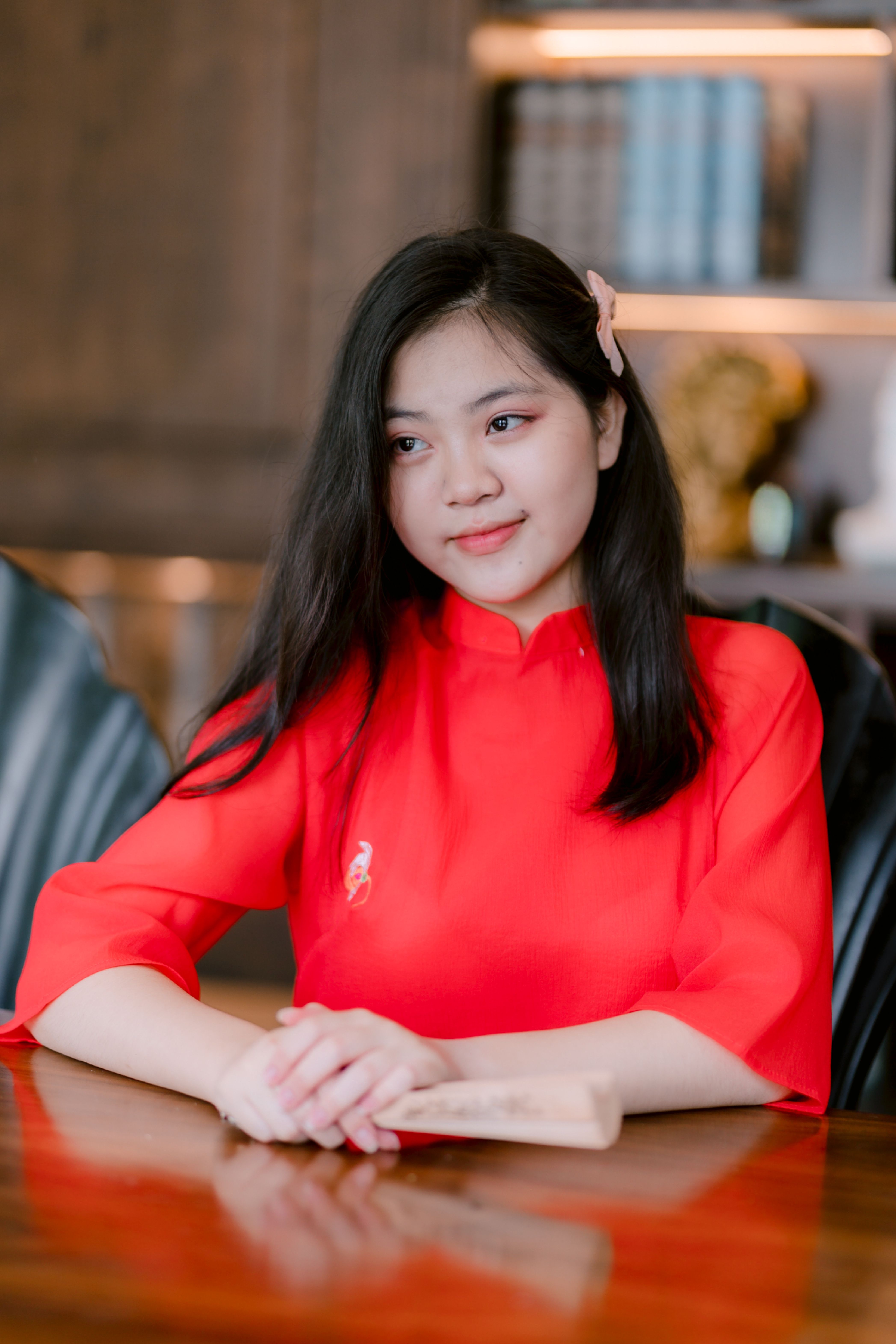 A young woman in a vibrant red dress sits at a table indoors, looking thoughtful.