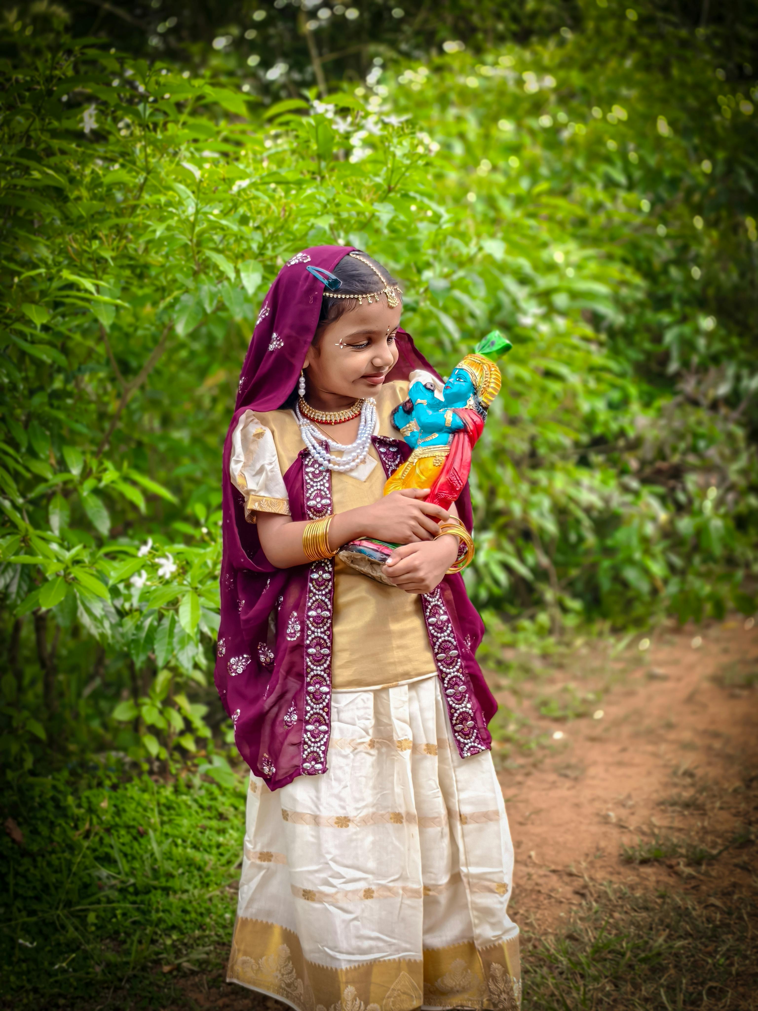 A young girl in vibrant traditional Indian attire holds a Lord Krishna idol outdoors, showcasing cultural heritage.