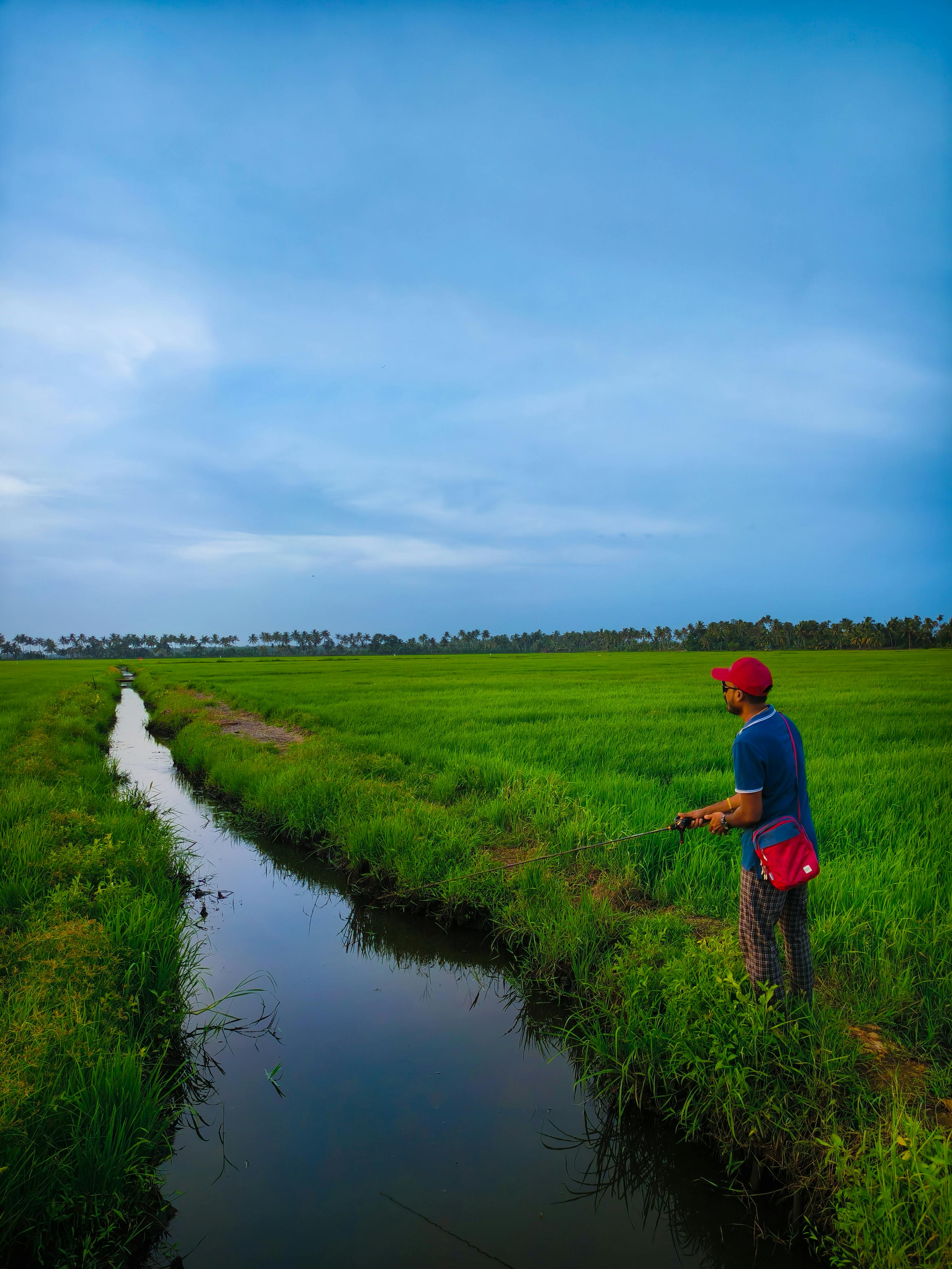 A man fishes along a canal in a vibrant green field under a bright blue sky at sunrise.