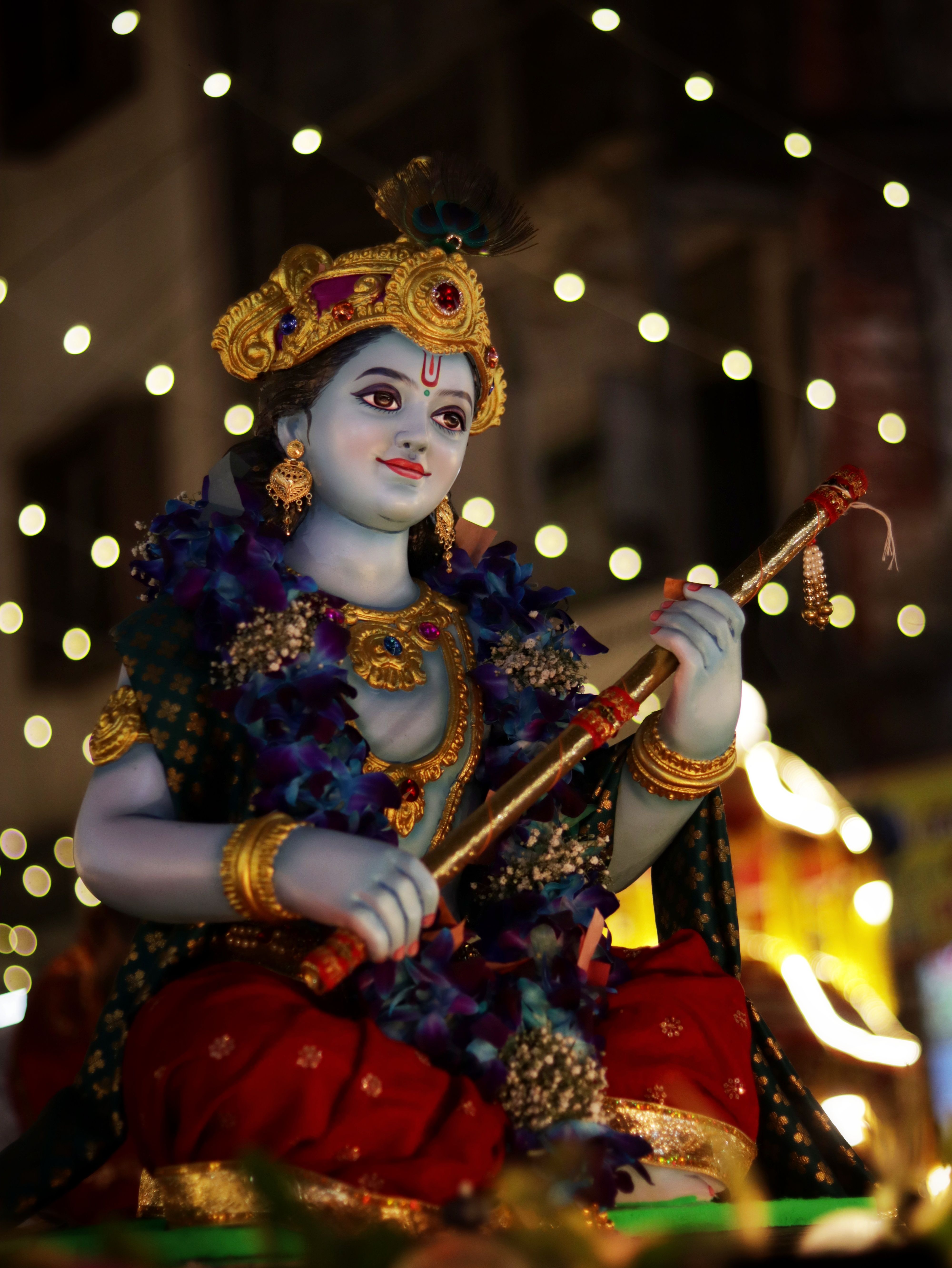 Colorful Krishna statue adorned with flowers and lights during a Hindu festival at night outdoors.