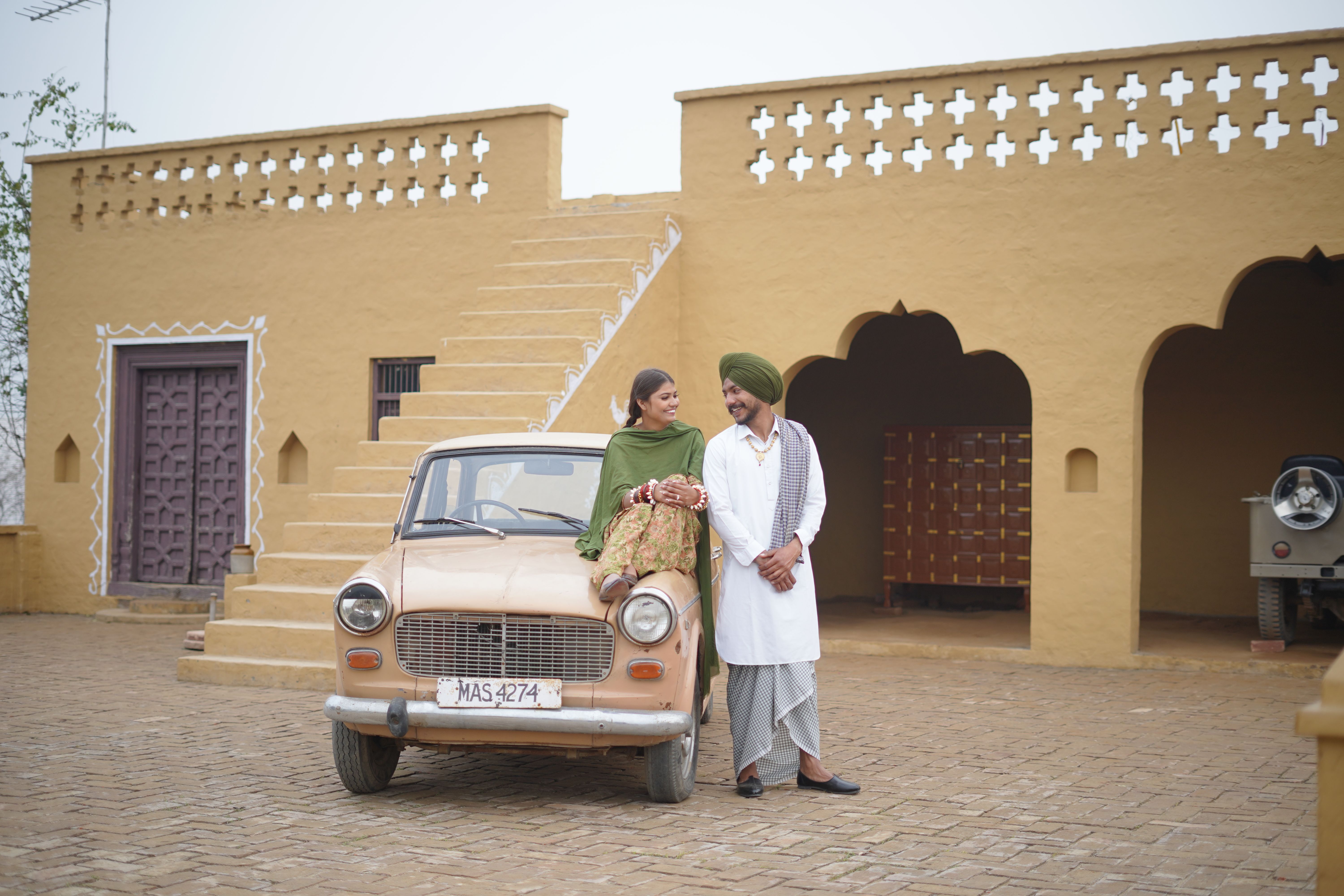 A couple in traditional attire enjoying a moment with a vintage car in a rustic courtyard.