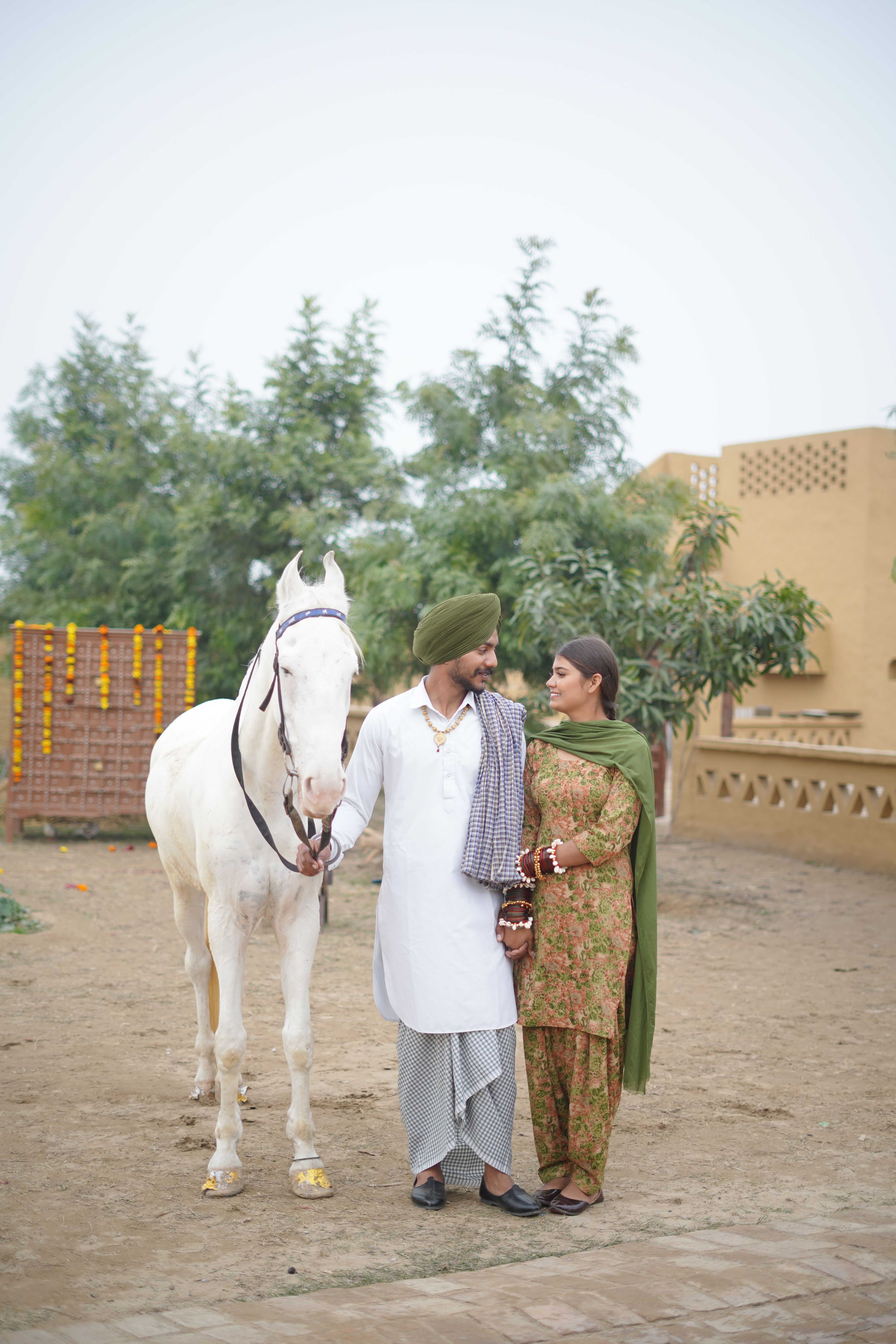 A couple dressed in traditional attire walks with a white horse in a rustic outdoor setting.