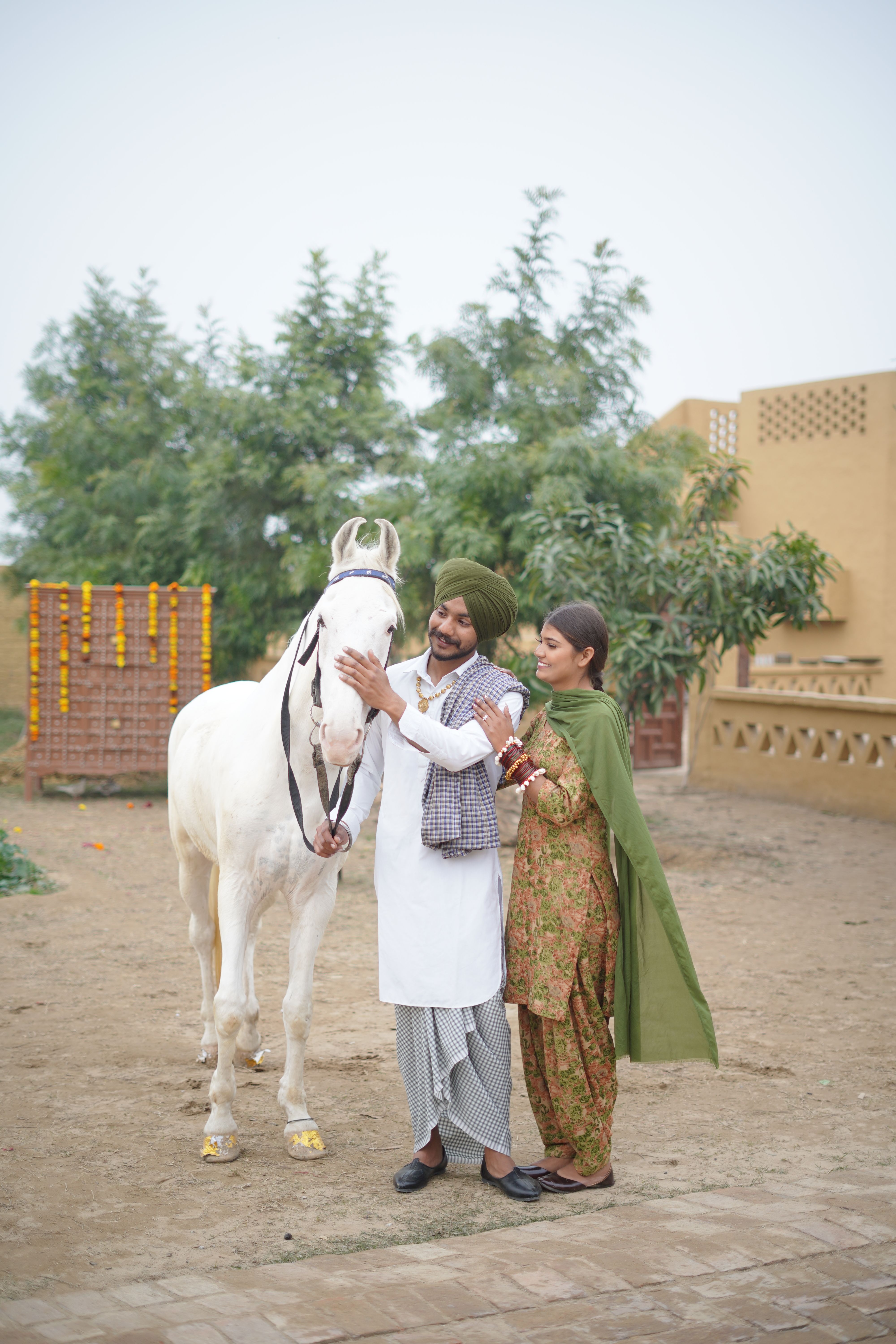 A couple in traditional attire lovingly embraces a white horse in an outdoor setting.
