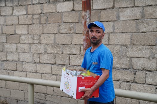 A male delivery worker in blue attire holding a packaged box outdoors.