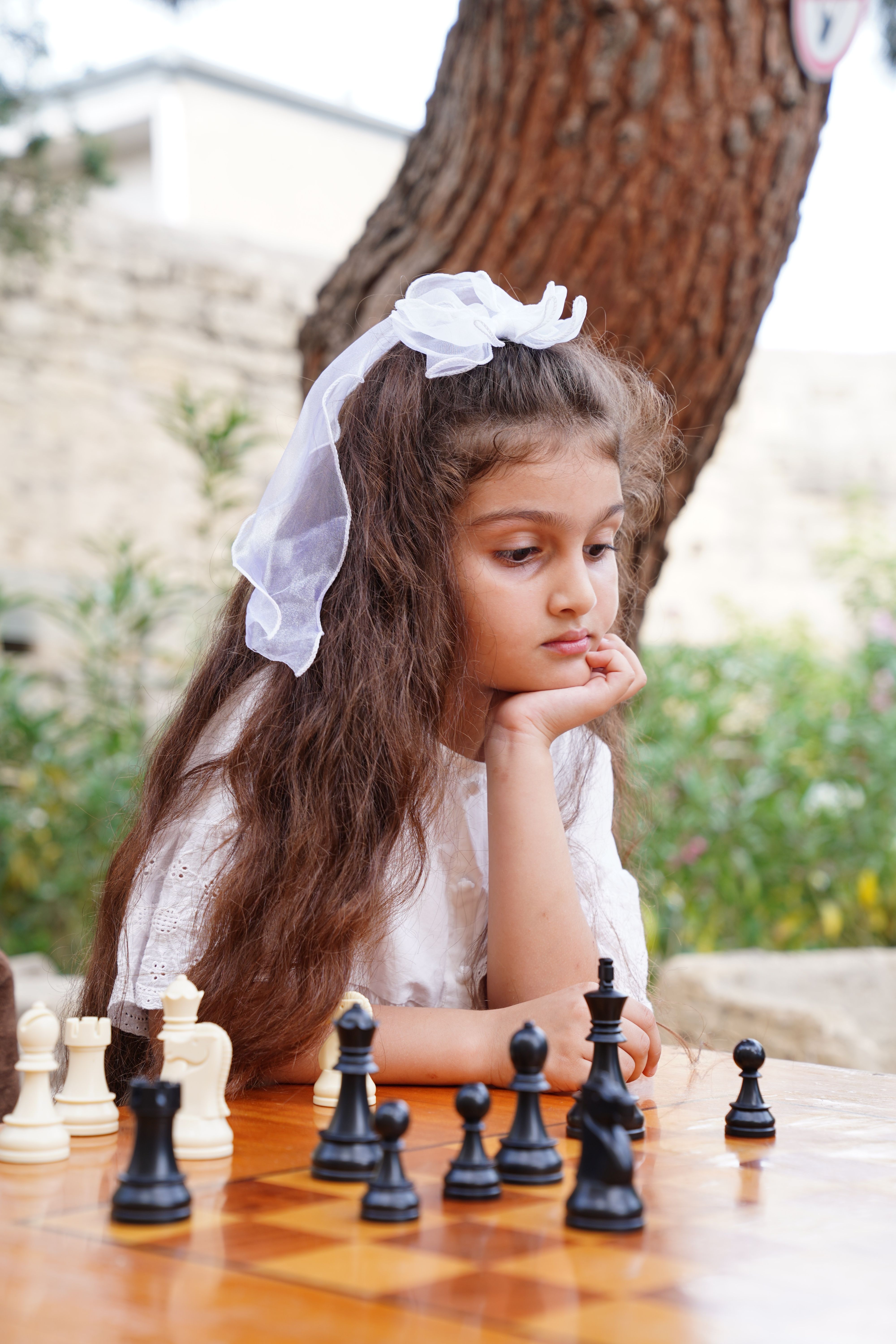 A young girl contemplates her next move in a chess game outdoors in a garden setting.