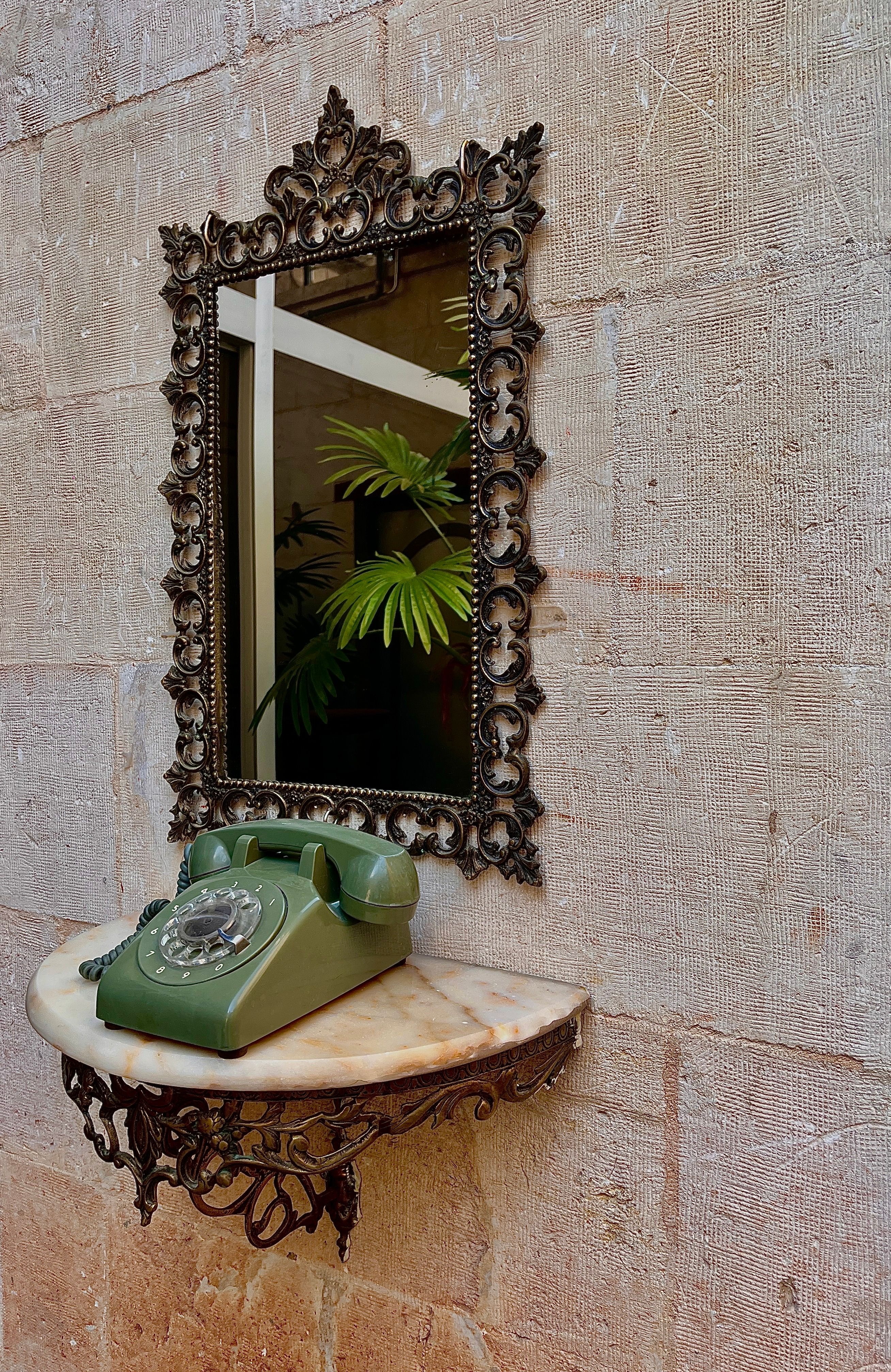 A vintage green rotary phone on a marble shelf with a decorative mirror.