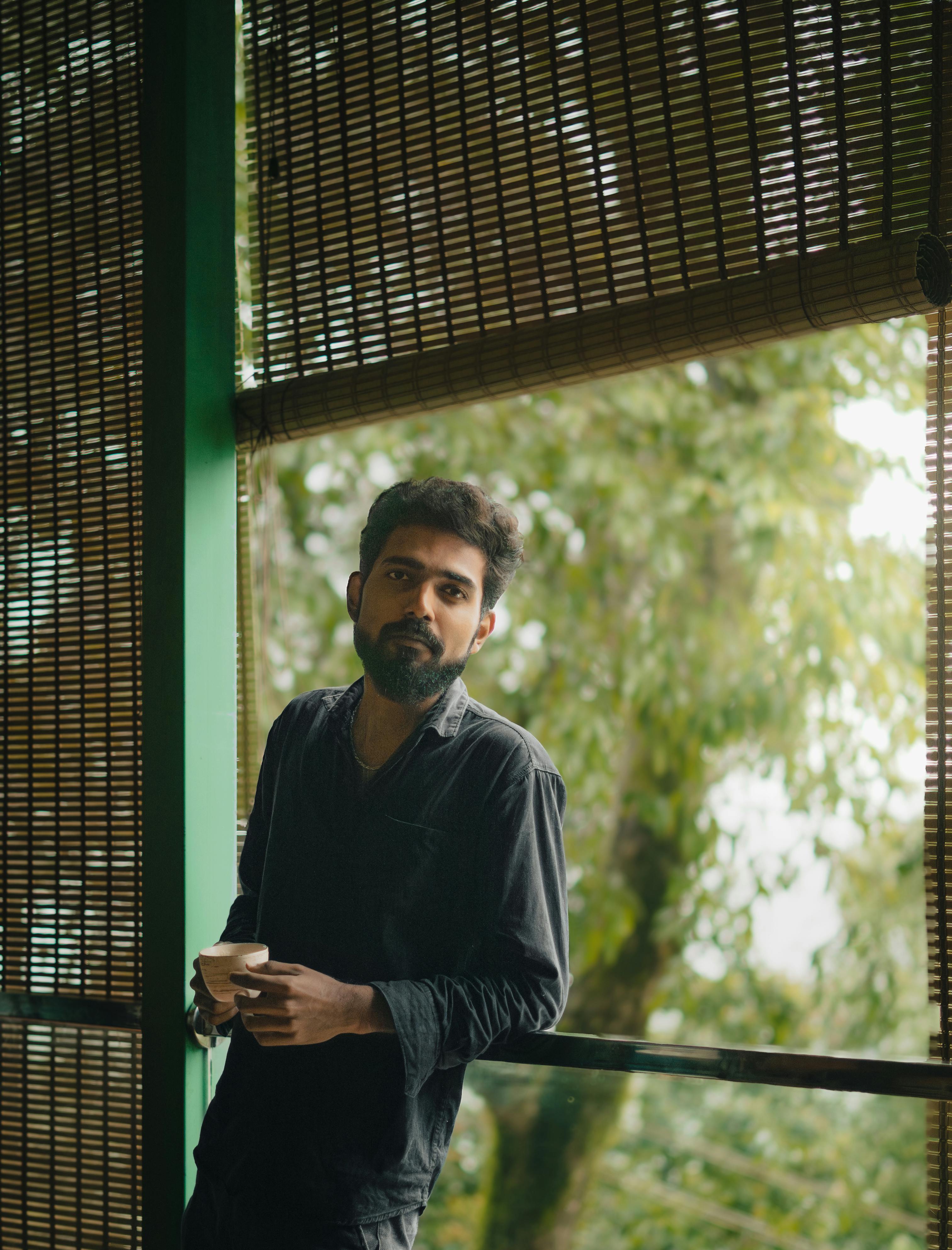 Man standing beside window, holding coffee cup, in relaxed indoor setting with greenery outside.