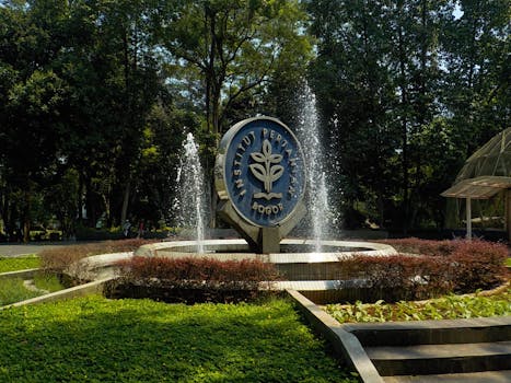 Fountain with the Institut Pertanian Bogor emblem surrounded by vibrant greenery and trees.