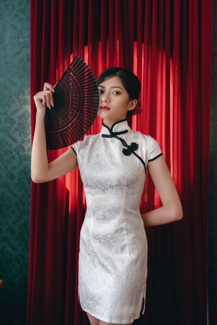 Adult woman in white qipao holding a red fan in front of a curtain, exuding elegance and tradition.