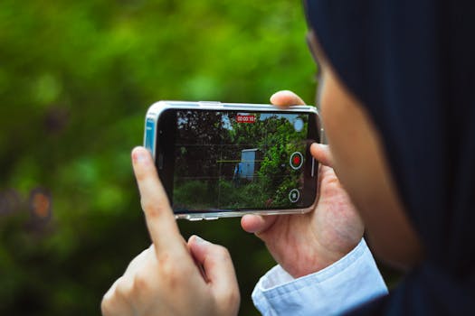 Close-up of person using smartphone to film a green outdoor scene in daylight.
