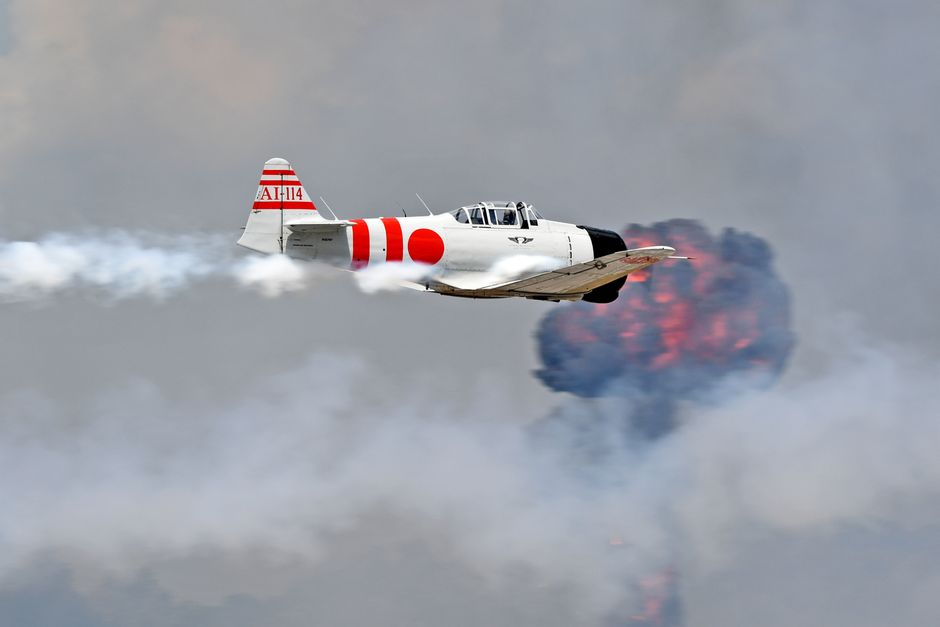 Vintage WWII Mitsubishi Zero aircraft with smoke trail flying against explosion backdrop.