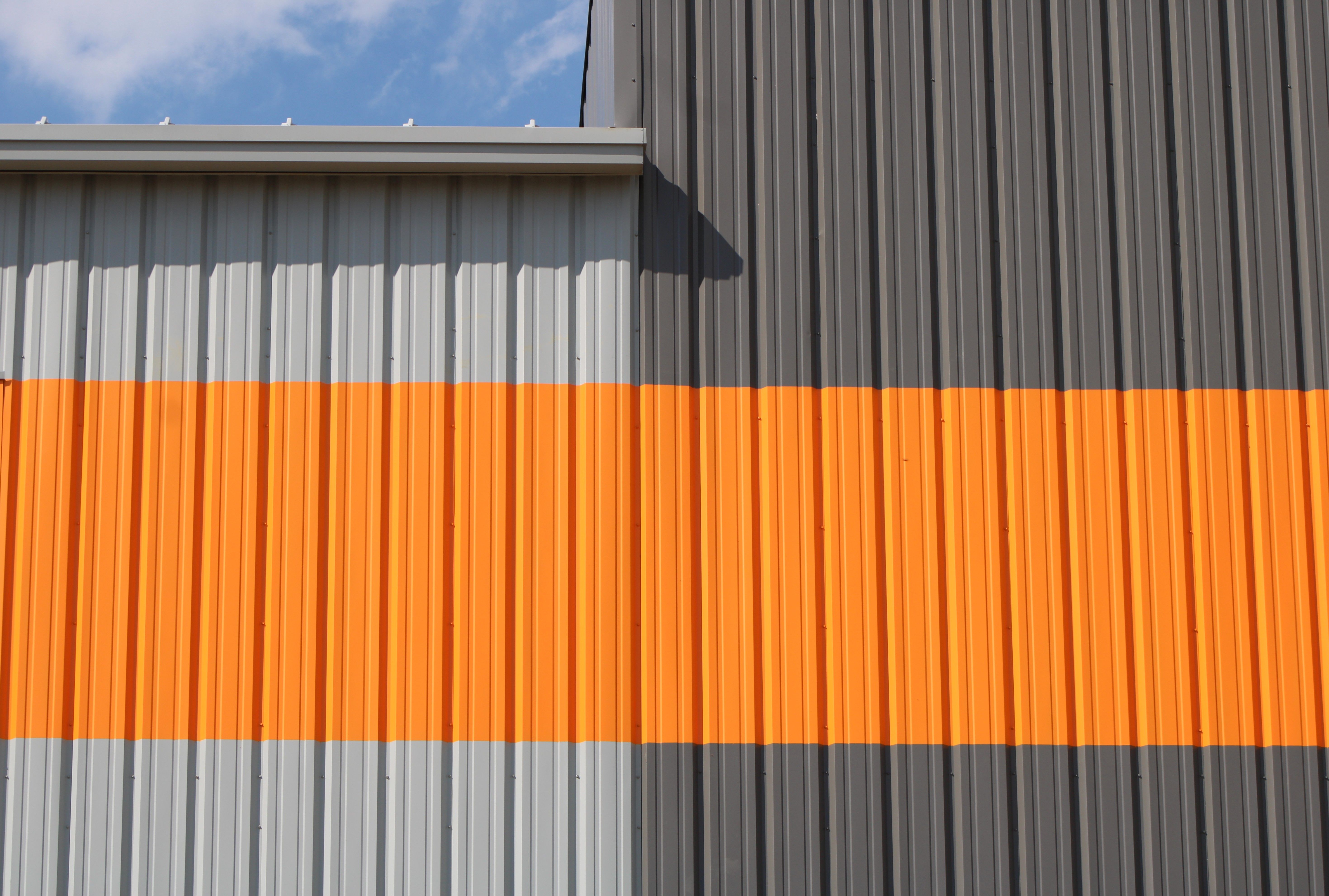 Close-up of industrial building facade with bright orange stripe against gray siding under blue sky.