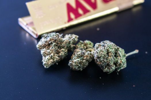 Macro image of cannabis buds accompanied by rolling paper on a dark surface.