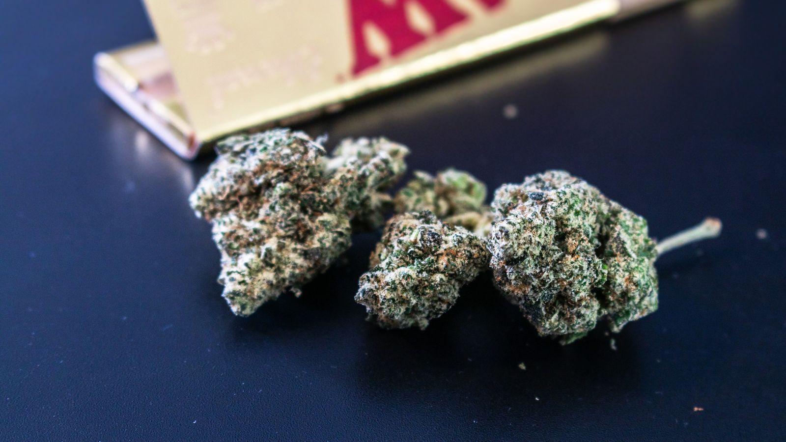 Macro image of cannabis buds accompanied by rolling paper on a dark surface.