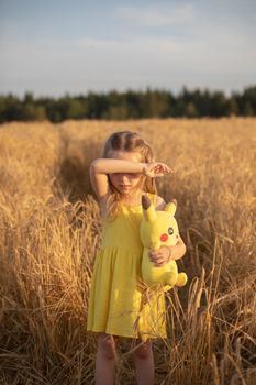 A young girl with a plush toy standing in a golden wheat field during sunset, covering her eyes.