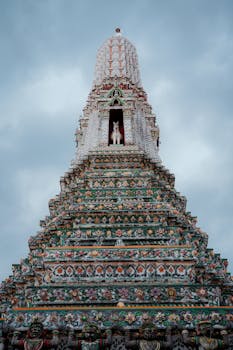 A detailed view of the ornate structure of Wat Arun temple under a cloudy sky in Bangkok, Thailand.