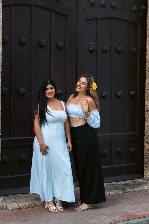 Free Two women stand smiling in front of a large wooden gate, embodying a lively outdoor summer vibe. Stock Photo