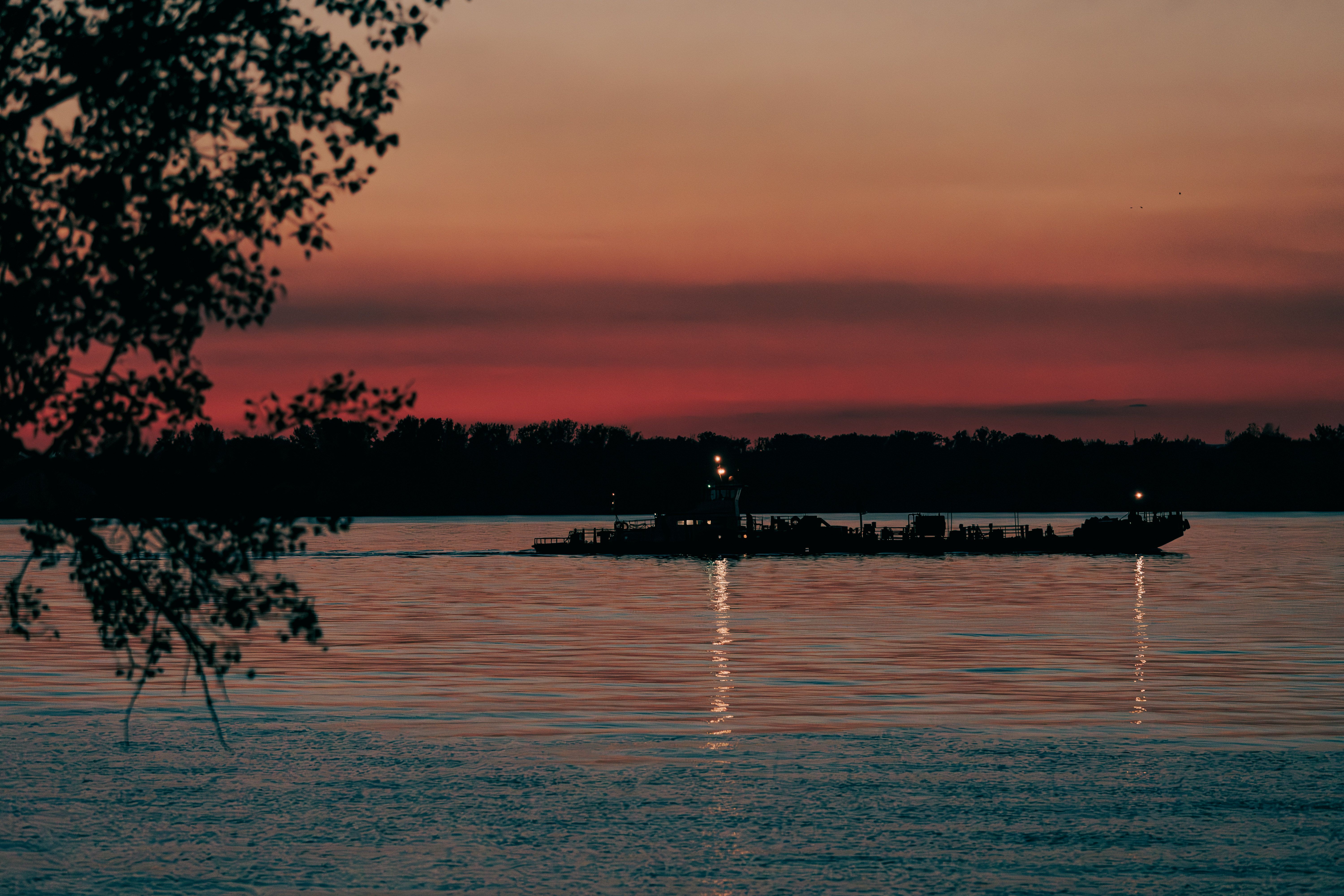A barge silhouetted on a wide river at dusk under a red sky, with tree branches in the foreground