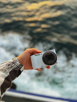 A hand holds a digital camera with ocean waves in the background, depicting travel and photography.